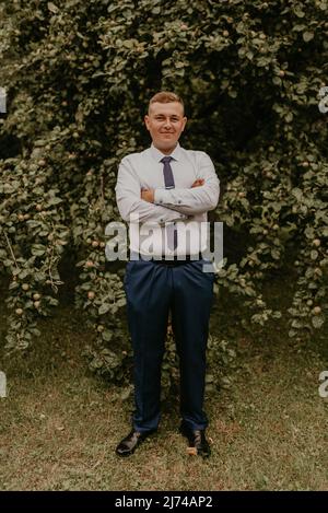 young blond man European curly-haired successful businessman groom in classic clothes white shirt and tie stands on street with his hands clasped Stock Photo