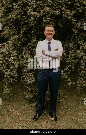 young blond man European curly-haired successful businessman groom in classic clothes white shirt and tie stands on street with his hands clasped Stock Photo