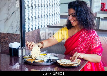 beautiful brunette woman in red sari eating with appetite traditional thali wirh rise,curd,dal in Goa restaurant masala tea Stock Photo