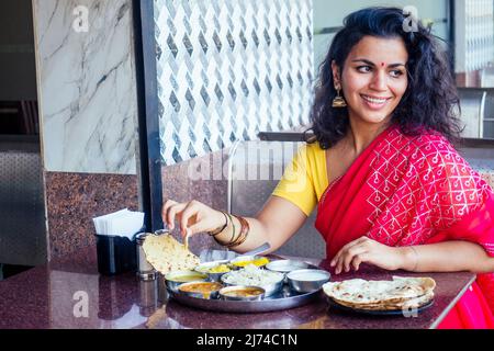 beautiful brunette woman in red sari eating with appetite traditional thali wirh rise,curd,dal in Goa restaurant masala tea Stock Photo