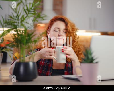 Portrait of young woman enjoying cup of coffee at home. Smiling beautiful lady drinking hot coffee at working on laptop. Excited female in plaid shirt laughs on autumn cloudy day. Stock Photo