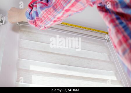 A handywoman with a tape measure installs white roller blinds indoors. Shutters day and night Stock Photo