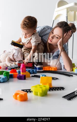 tired mother lying near toddler son playing with toy train on white Stock Photo