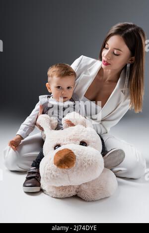 baby boy looking at camera while sitting on toy dog near mother on grey Stock Photo