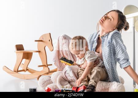 tired woman sitting near playing son and rocking horse on white Stock Photo