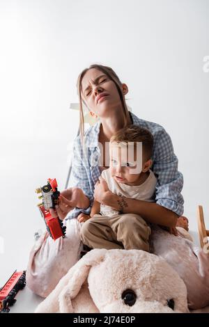 tired woman with closed eyes embracing son and holding toy train on white Stock Photo