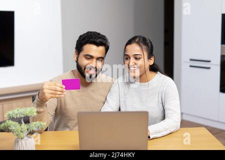 Portrait of a smiling young multiracial couple shopping online while sitting on a new modern kitchen with credit card and laptop computer, buying furniture for a new apartment Stock Photo