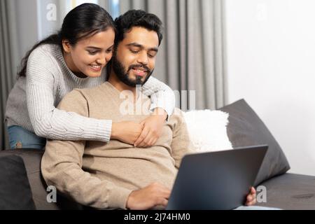 Cheerful couple in love spending leisure time online with a laptop at home. Young woman and man looking at the laptop screen while sitting on the sofa, watching comedy movies, funny videos Stock Photo