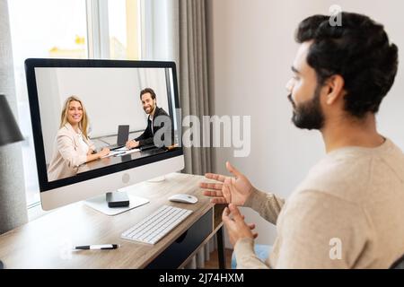 Serious concentrated pakistani male student involved virtual meeting, webinar. Man making video call on the computer, sitting at the table at home, talking and gesturing, explaining smth Stock Photo