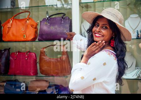 happy indian woman wearing straw hat and standind near showcase with fashion bags in India Goa market Stock Photo