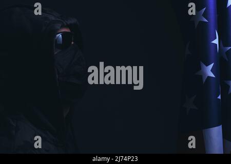 Hooded man wearing black glasses and mask looks flag of the United States America.Hacker attacker and bandit Stock Photo