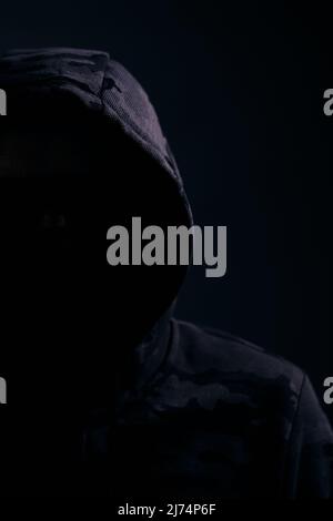 Man in dark camouflage hoodie with covered face dark background, hacker bandit and attacker Stock Photo