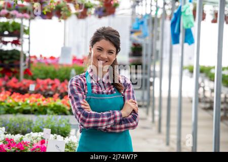 Portrait of pretty woman with apron standing with crossed arms in plant nursery centrum in springtime with different flowers in background Stock Photo