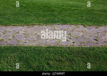 garden path made of natural stone paved with rough rock overgrown with grass in a park with a green lawn close-up of the path with an abstract pattern Stock Photo