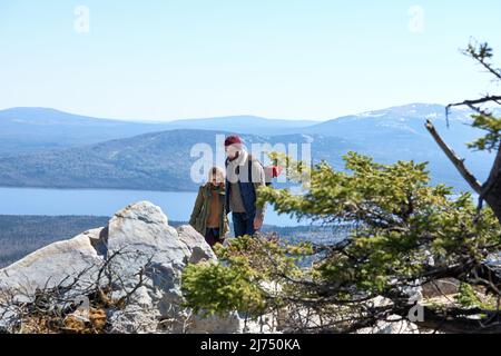 Young couple with backpacks climbing up the rocks on scenic background of wide river, green hills and snow-capped mountains Stock Photo