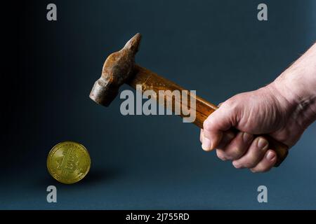 A man hits a bitcoin coin with an old rusty hammer. Male hand with a hammer and a bitcoin coin close-up on a gray background. Stock Photo