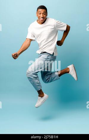 Lucky joyful African-American young guy jump up on blue background, having fun in studio, cheerful man wearing casual t-shirt and jeans celebrating good news, victory, jumping for happiness, vertical Stock Photo