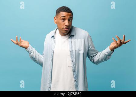 Confused multiracial guy in casual shirt shrugging shoulders isolated on blue background, uncertain African-American man feels doubt, hesitant man in bewilderment spreads palms Stock Photo