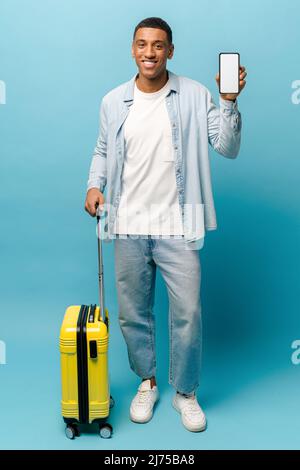 Full length portrait of cheerful young African-American guy in casual wear stands with yellow suitcase and showing phone with empty screen, isolated on blue, man advertising mobile app for travel Stock Photo