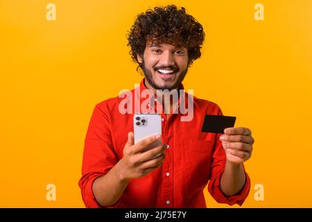 Middle eastern happy man is ordering food online, male paying for long-awaited purchase on the mobile phone. Guy is using smartphone and credit card for shopping online isolated on orange Stock Photo