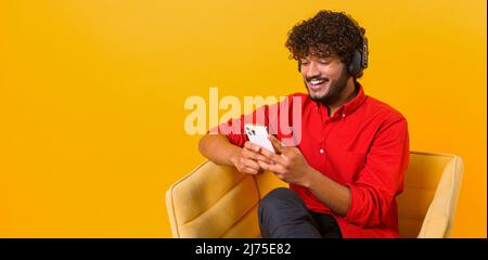 Studio portrait of young middle eastern guy in wireless headphones isolated on orange wall, looking at mobile phone, enjoying listening to favorite music soundtrack Stock Photo