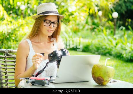 Female photographer inserting or removing a memory card in her professional dslr camera as she sits at a desk with her laptop at paradise beach green Stock Photo