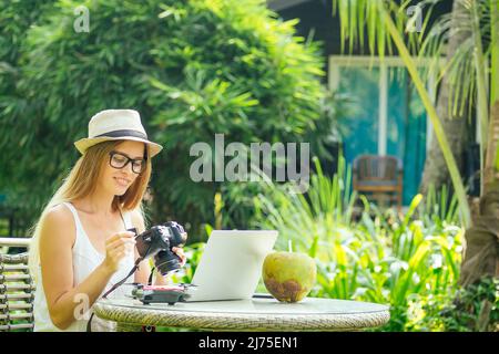 Female photographer inserting or removing a memory card in her professional dslr camera as she sits at a desk with her laptop at paradise beach green Stock Photo