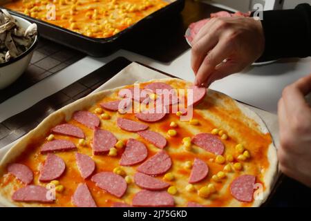 Man cooking homemade pizza on a wooden table in a home kitchen. Male hand make pizza dough with red tomato sauce. Base of Margarita. Home culinary Stock Photo