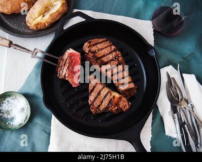 Two Grass Fed Steaks in a Cast Iron Skillet with Baked Potatoes in the Background Stock Photo