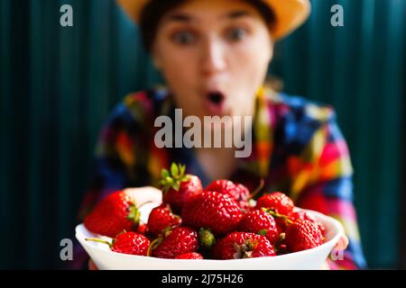 Defocus young woman in a hat and shirt smiles and shows a white bowl of strawberries. Dark green steel background. Summer food, fruits. Summertime Stock Photo