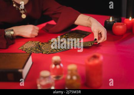 KYIV, UKRAINE - FEBRUARY 23, 2022: Cropped view of medium taking tarot card near blurred jars and book on table Stock Photo