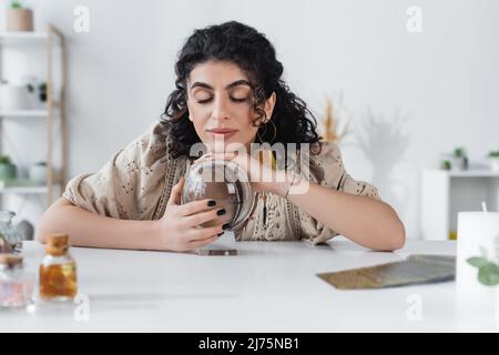 Young gypsy medium touching orb near blurred jars and tarot cards on table Stock Photo