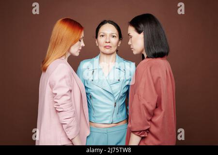 Serious mature mother looking at camera and standing between multi-ethnic daughters against brown background Stock Photo