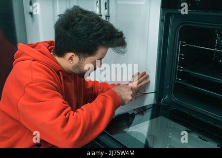 Repairman, young mechanic fixes a broken built-in oven, man using screwdriver tightening screws, self repair Stock Photo