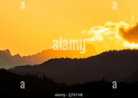 Sunset on Germany's highest mountain, the Zugspitze in gold & red with black forest in the foreground. A cloud pushes from the right into the picture. As background taken from the shore of Walchensee. Stock Photo