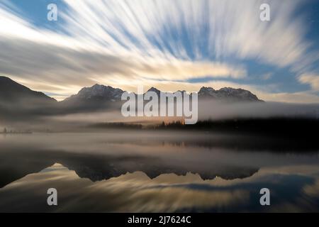 Nighttime long exposure of the clouds and fog in autumn, with the Karwendel mountains reflected in the clear water of the Barmsee near Krün. Stock Photo