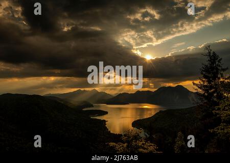 Sun rays and golden light over Walchensee, with Herzogstand, Heimgarten and Simetsberg in the background. The lake reflects the color and dense clouds in the sky. Stock Photo