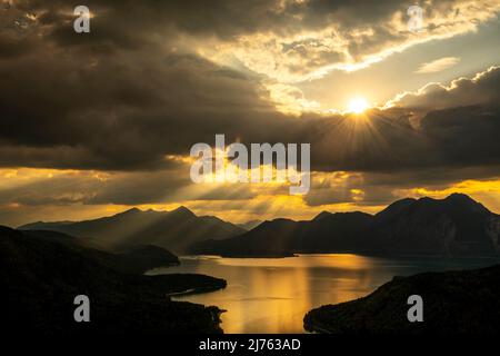 Sun rays and golden light over Walchensee, with Herzogstand, Heimgarten and Simetsberg in the background. The lake reflects the color and dense clouds in the sky. Stock Photo