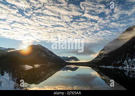 The evening sun in winter on the shore of the Sylvenstein reservoir. Inetressant clouds and fog atmosphere as a reflection in the clear water of the alpine lake in the Bavarian Alps on the edge of the Karwendel. Stock Photo