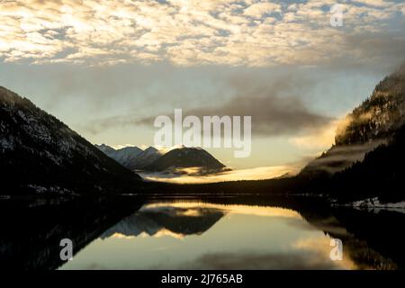 Fog and cloud atmosphere at the Sylvensteinspeicher, a lake / reservoir in the Bavarian Alps on the edge of the Karwendel. The evening light mood is reflected in the water of the lake and differently colored clouds or fog banks give the scenery something mystical. Stock Photo