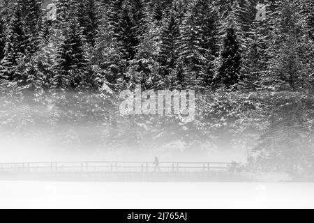 Three walkers on the snowed-in bridge at the outlet of the Lautersee in the Bavarian Alps in winter with fresh snow and dense ground fog Stock Photo