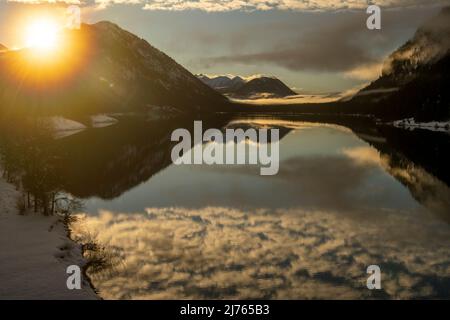 The evening sun in winter on the shore of the Sylvenstein reservoir. Inetressant clouds and fog atmosphere as a reflection in the clear water of the alpine lake in the Bavarian Alps on the edge of the Karwendel. Stock Photo