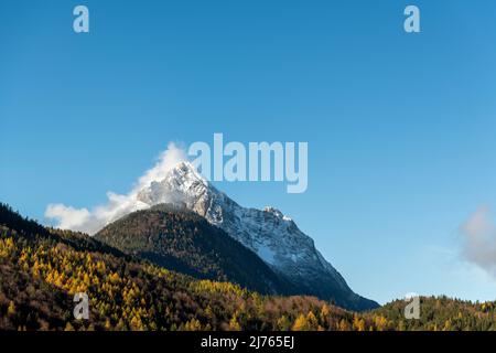 The Wetterstein, part of the mountain range of the same name between Garmisch-Partenkirchen and Mittenwald, seen from the village in autumn leaves and blue sky, with snow on the mountain. Stock Photo