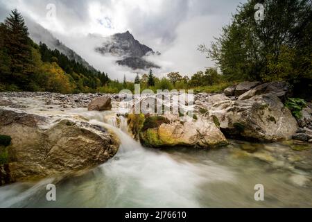 Dense clouds and fog briefly give the view of the Spritzkarspitze of the Lalidererwände at the Großer Ahornboden near Hinterriss in Tyrol, Austria in the Karwendel. In the foreground the Rissbach of the Engtal, with a small waterfall between large boulders. Stock Photo