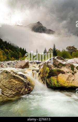 Dense clouds and fog briefly give the view of the Spritzkarspitze of the Lalidererwände at the Großer Ahornboden near Hinterriss in Tyrol, Austria in the Karwendel. In the foreground the Rissbach of the Engtal, with a small waterfall between large boulders. Stock Photo