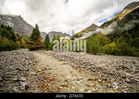 Dense clouds and fog briefly give the view of the Spritzkarspitze of the Lalidererwände at the Großer Ahornboden near Hinterriss in Tyrol, Austria in the Karwendel. In the foreground the Rissbach of the Engtal, in its streambed. Stock Photo