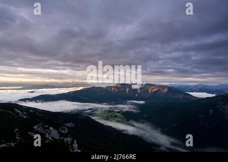 The so-called mountain 'Unnütz' and the underlying village 'Steinberg im Rofan' during light alpenglow in the early morning at sunrise seen from the Guffert, fog rolling along the valley. Stock Photo