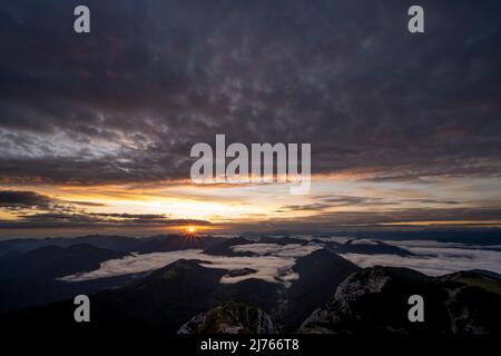 Sunrise with sun star and light colored clouds, while in the valley fog and clouds are moving. View from Guffert (2194m) in Rofan early in the morning. Stock Photo