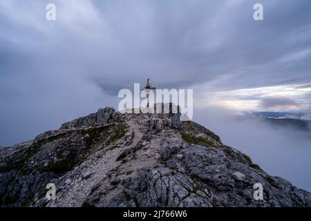 The path to the summit and summit cross of the Guffert in bad weather and strong wind during sunset. Dense clouds and fog obscure much of the colors, but a slight colored glow from the setting sun shines through. Stock Photo