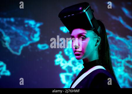 Portrait of serious lady gamer with virtual reality headset standing against digital world map background Stock Photo